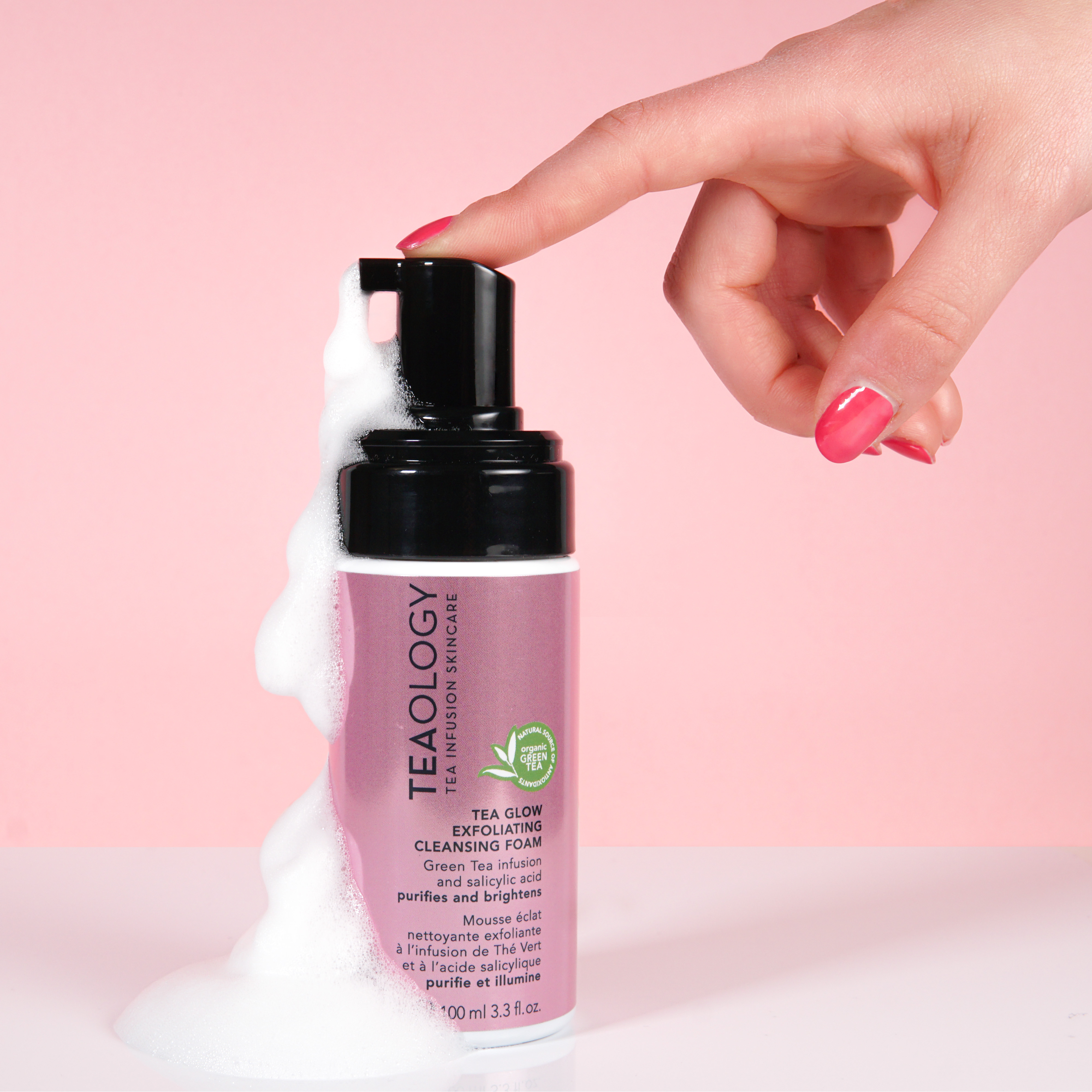 Hand drückt Schaum aus der Tealogy Tea Glow Exfoliating Cleansing Foam Flasche vor rosa Hintergrund.